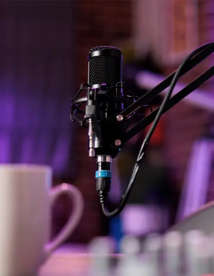 Black condenser microphone on a boom arm shock mount in a purple-lit studio setup with a mug in the foreground.