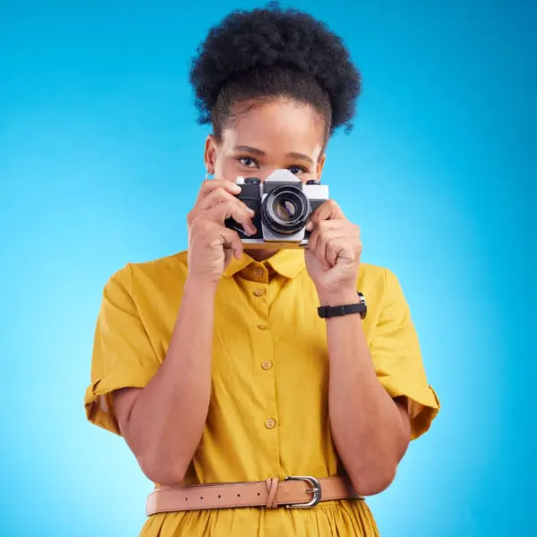 Woman in a yellow dress holding a vintage film camera up to her eye against a bright blue background.