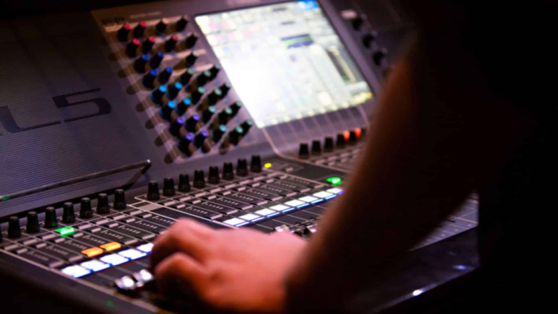 Hand operating an Allen & Heath digital mixing console with glowing buttons and built-in touchscreen display.