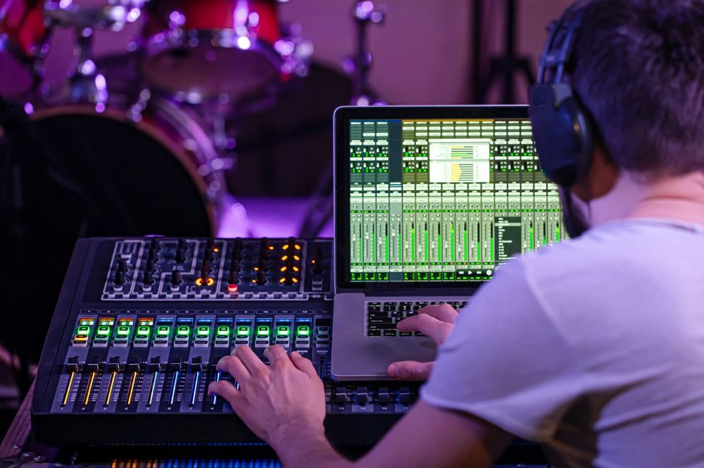 Sound engineer with headphones operating a digital mixing console and laptop DAW, with drums in background.