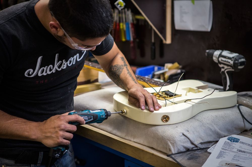 Tattooed guitar tech in a Jackson shirt using a power drill to assemble a cream electric guitar body in a workshop.