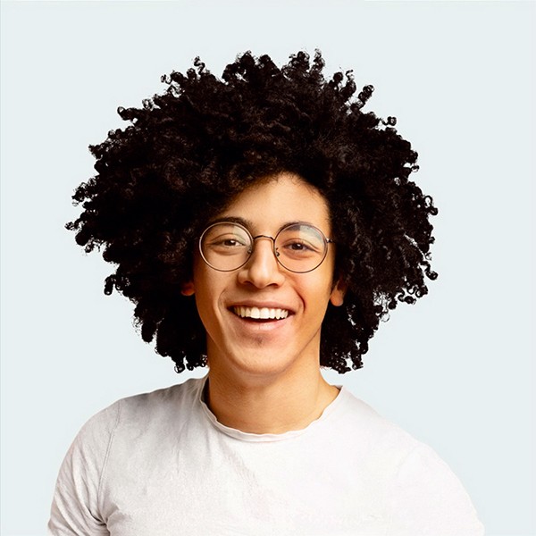 Smiling young man with a large curly afro and round glasses wearing a white t-shirt against a light background.