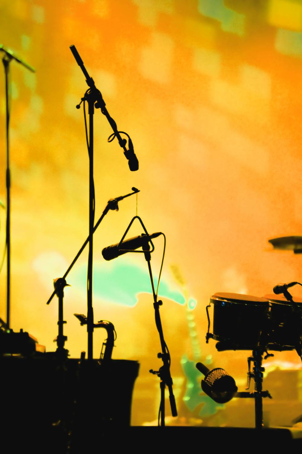 Silhouettes of microphones on stands and a drum kit against vibrant orange and green stage lighting at a live concert.