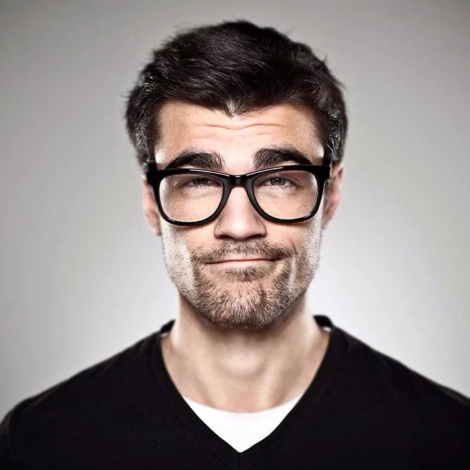 Man with dark hair, thick black glasses, and a beard smiling in a professional headshot against a grey background