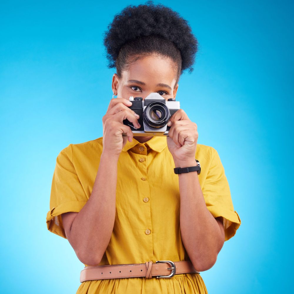 Woman in a yellow dress holding a vintage film camera up to her eye against a bright blue background.