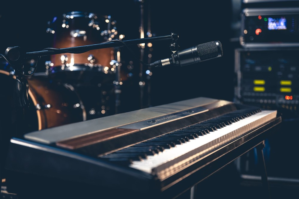Electric keyboard with condenser microphone on a boom stand in a dimly lit studio, drums and rack gear in background.