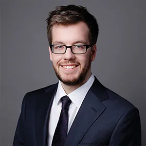 Young man with glasses and a beard in a navy suit and tie, smiling in a professional headshot on a grey background.