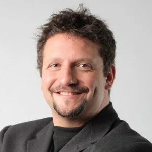 Man with dark curly hair and a goatee in a black blazer, smiling in a professional headshot on a grey background.