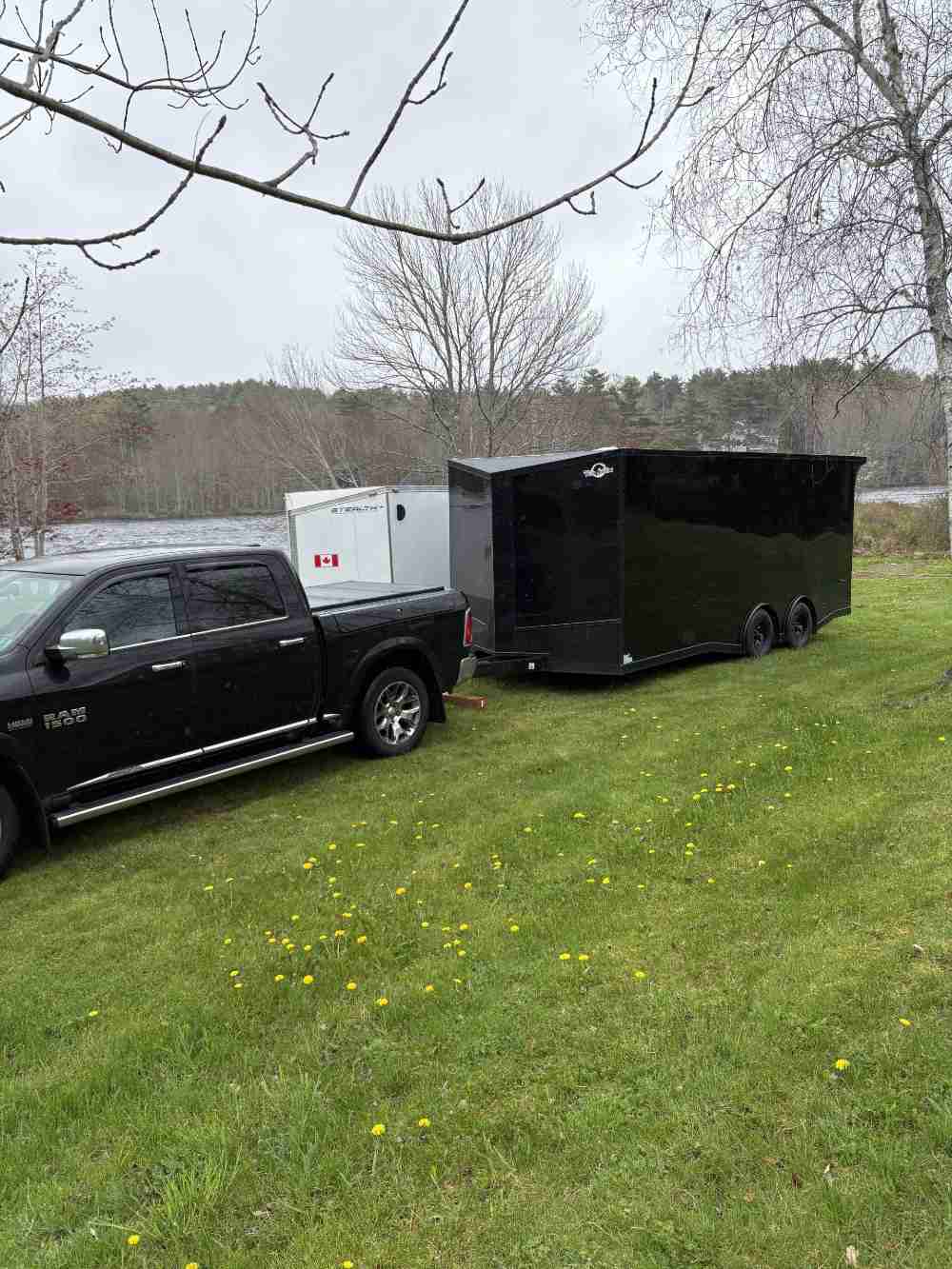 Black Ram 1500 pickup truck towing a large black enclosed cargo trailer on a grassy field beside a lake in Canada.