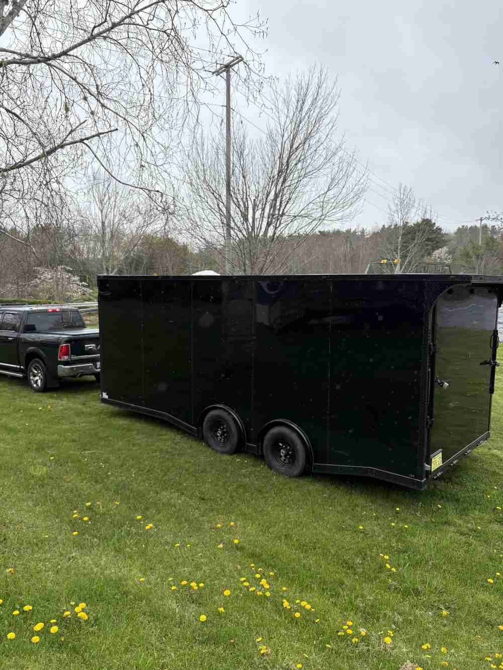 Large black enclosed cargo trailer with dual axles parked on a grass field, with a black Ram truck visible behind it.
